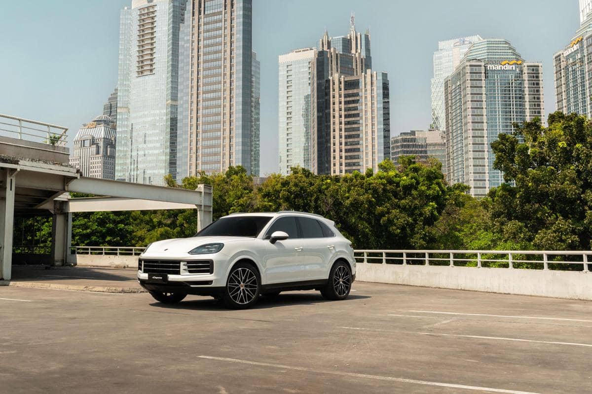 SUV Porsche Cayenne berwarna putih diparkir di area terbuka dengan latar gedung pencakar langit modern di siang hari.