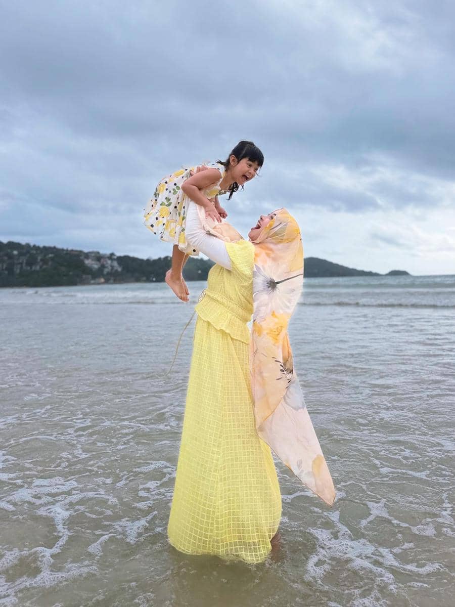 Seorang wanita berhijab berwarna kuning mengangkat anak kecil di tepi pantai Phuket dengan latar langit mendung dan ombak tenang.