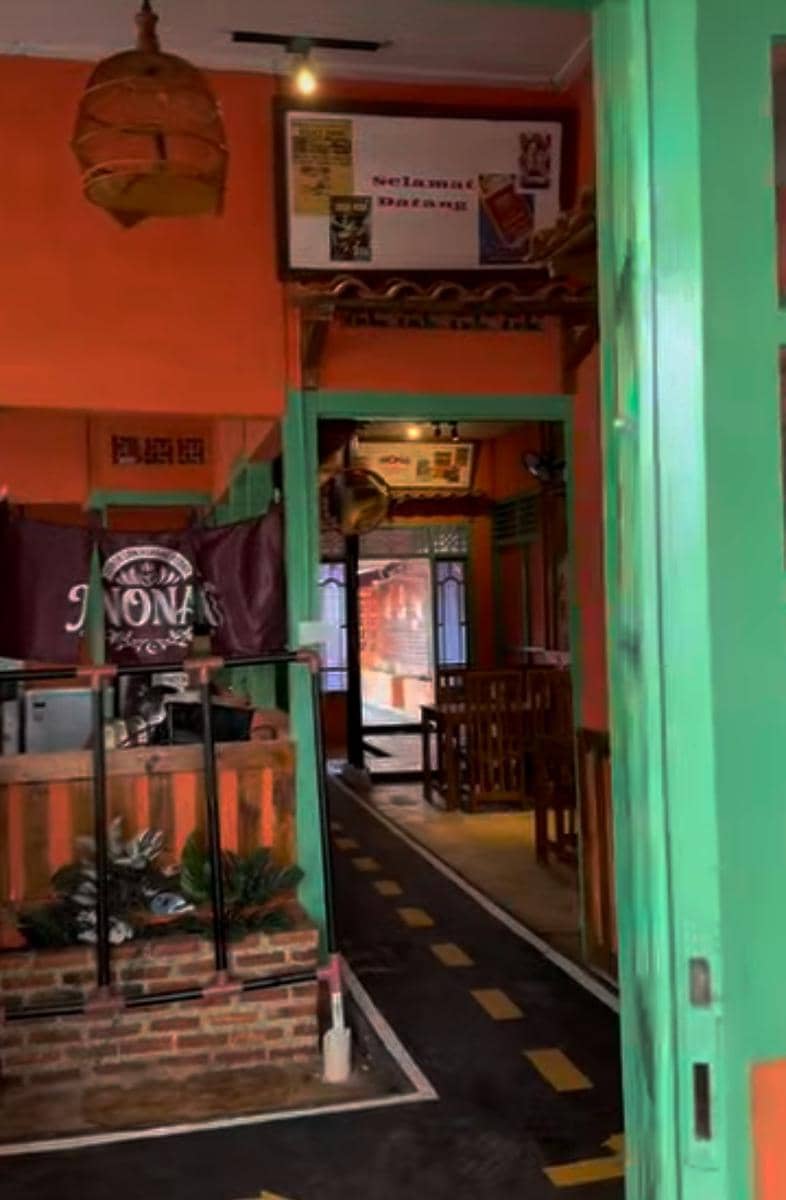 Interior warung Ceker Kerang Pedas Nona di Bandar Lampung dengan dinding oranye, pintu hijau, dan meja kayu untuk pengunjung.