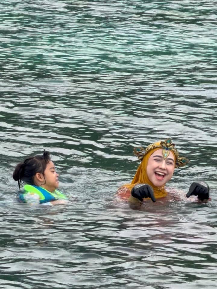 Seorang perempuan berhijab mengenakan mahkota berenang di laut bersama seorang anak kecil yang memakai pelampung berwarna hijau.