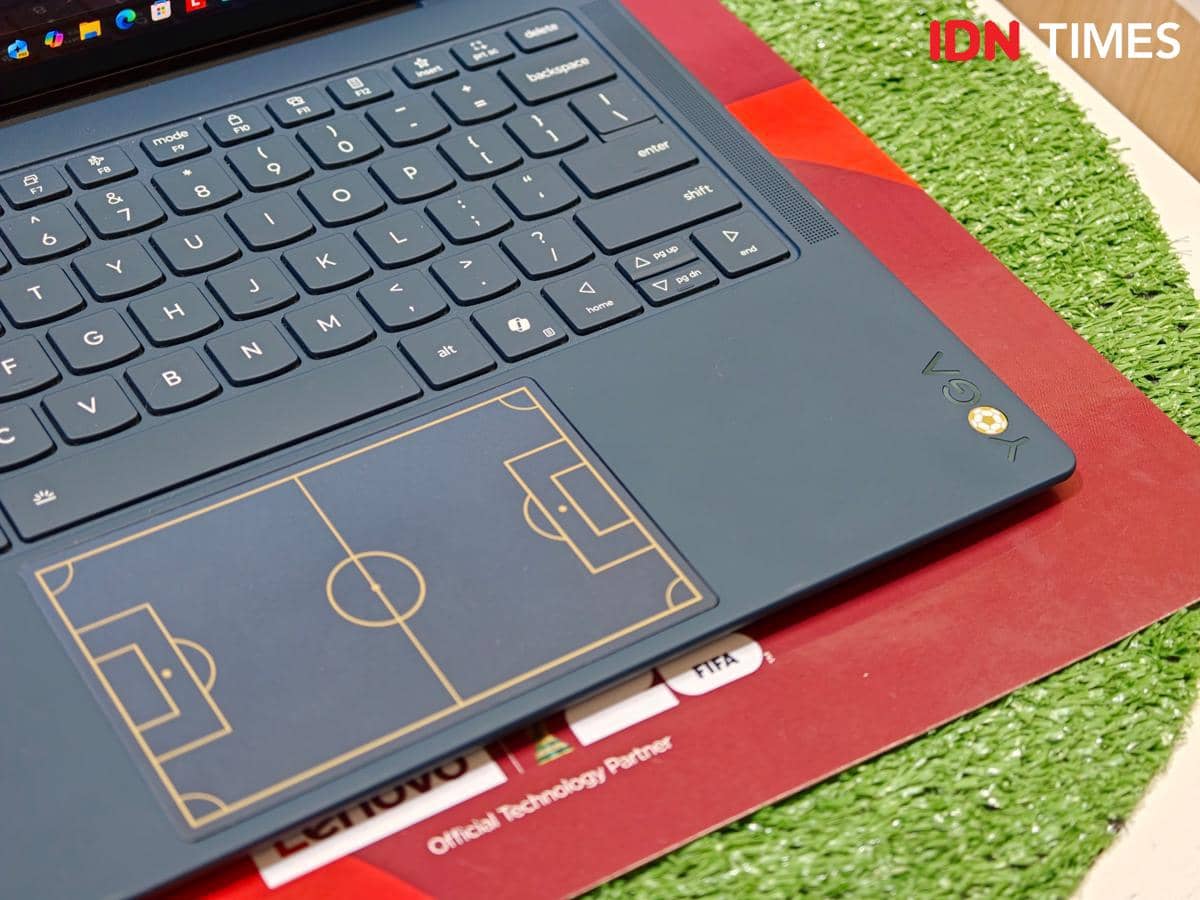 Keyboard laptop Lenovo dengan desain lapangan sepak bola pada touchpad, ditempatkan di atas alas berwarna merah dan hijau.
