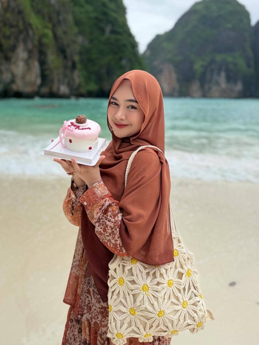 Seorang perempuan berhijab cokelat tersenyum sambil memegang kue ulang tahun kecil di tepi pantai dengan latar tebing hijau di Phuket.