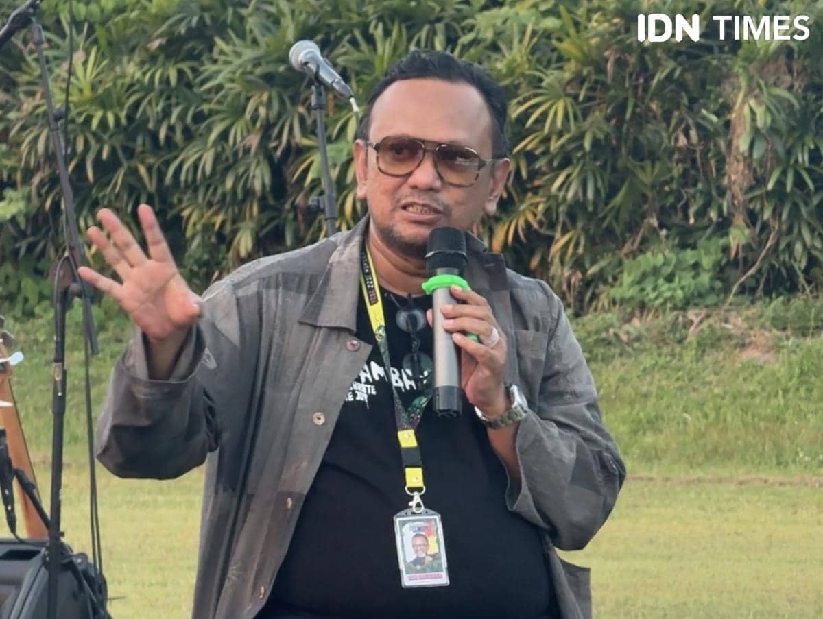 Seorang pria mengenakan kacamata dan jaket abu-abu memegang mikrofon sambil berbicara di luar ruangan dengan latar pepohonan.