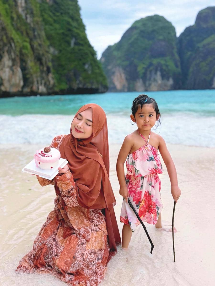 Seorang perempuan berhijab memegang kue ulang tahun kecil di tepi pantai bersama seorang anak kecil di air dangkal dengan latar tebing hijau Phuket.