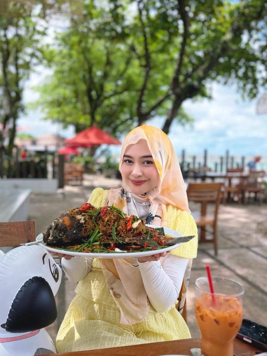 Seorang wanita berhijab mengenakan pakaian kuning duduk di restoran tepi pantai sambil memegang sepiring ikan goreng besar dengan sambal dan sayuran.