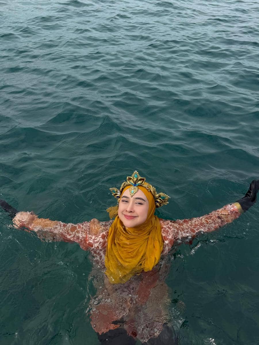 Seorang perempuan berhijab kuning dan mengenakan mahkota emas tersenyum sambil berada di air laut biru di Pantai Phuket.