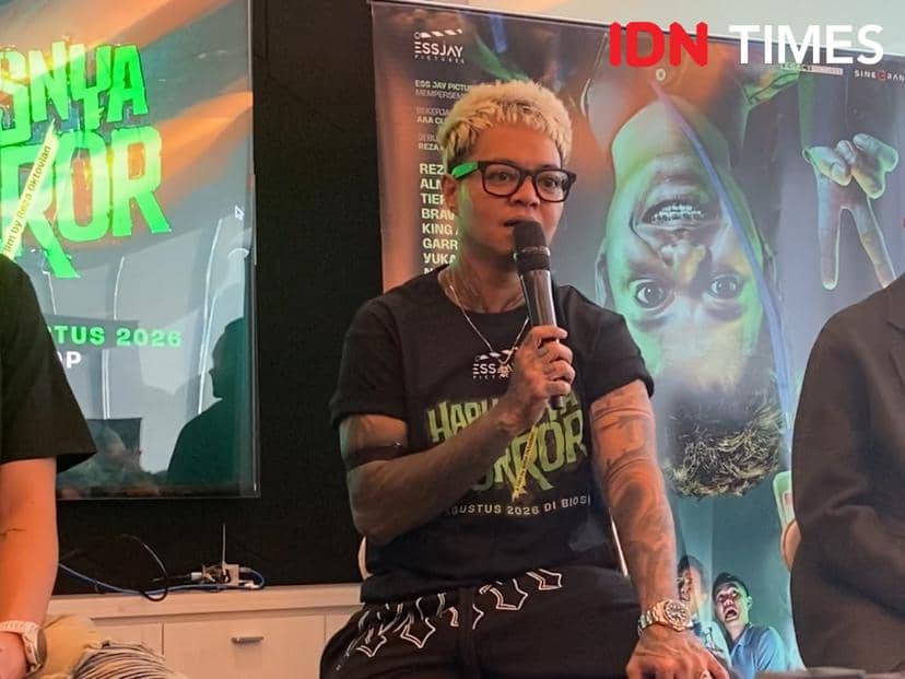 Seseorang berambut pirang pendek mengenakan kacamata hitam dan kaus hitam bertuliskan Harusnya Horor berbicara memegang mikrofon di acara promosi film.