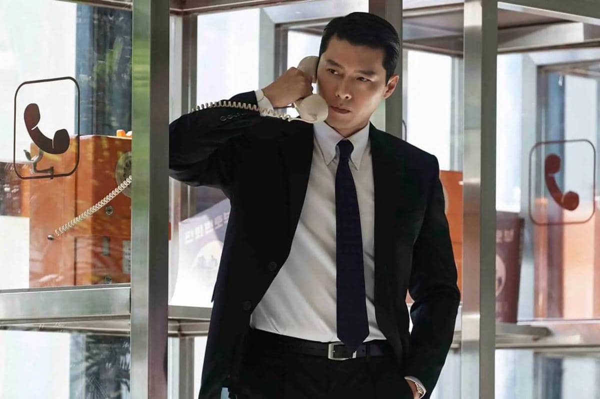 Seorang pria mengenakan setelan jas hitam sedang berbicara di telepon umum di dalam bilik kaca dengan ekspresi serius.