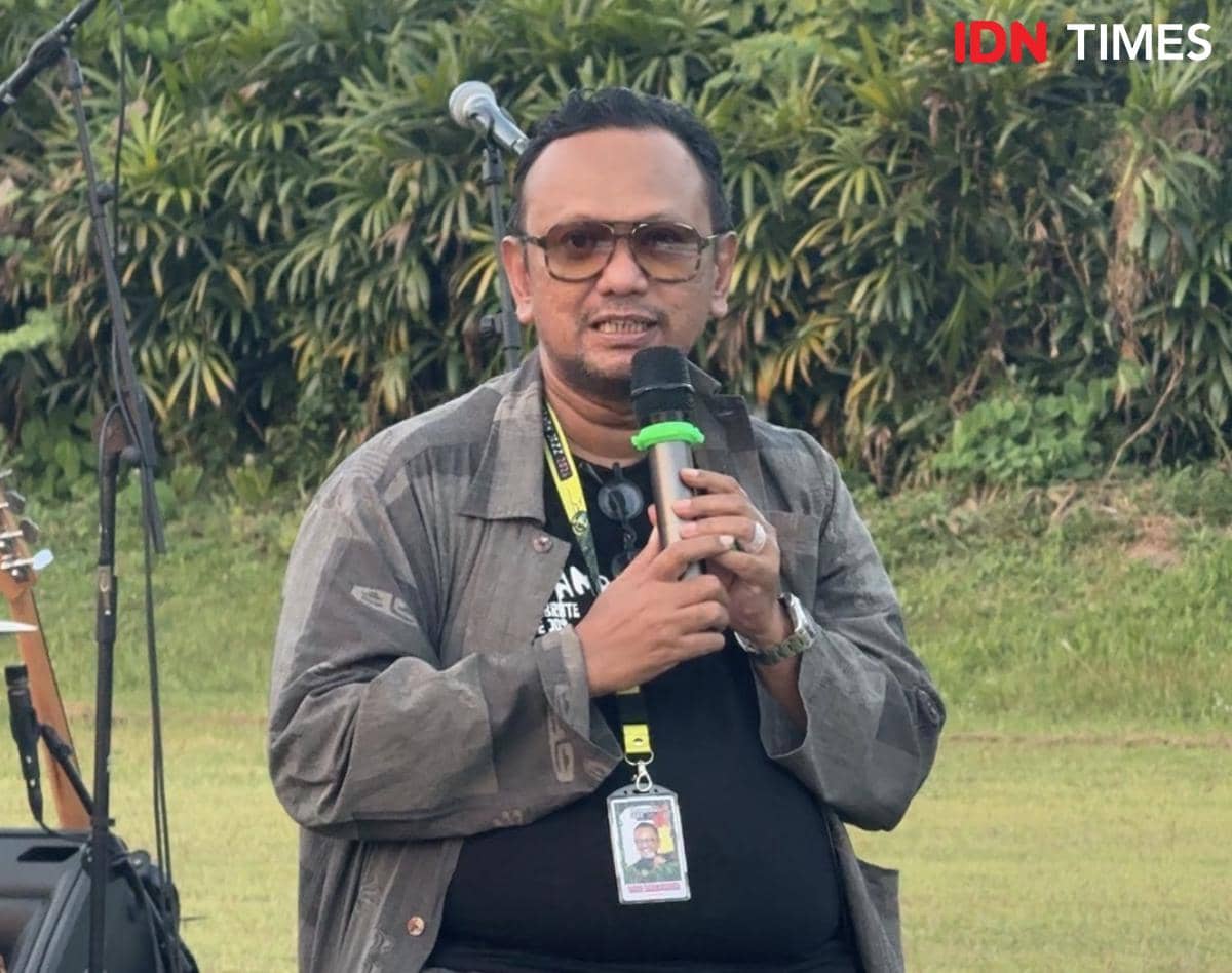 Seorang pria mengenakan kacamata dan jaket abu-abu berbicara memegang mikrofon di area luar ruangan dengan latar pepohonan.