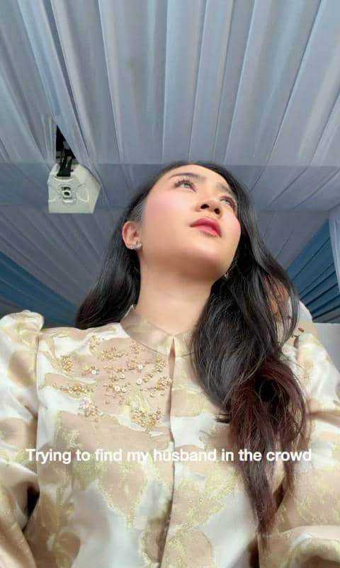 Seorang wanita berbusana formal berwarna krem dengan hiasan bordir emas menatap ke arah atas di bawah tenda berwarna putih dan biru.