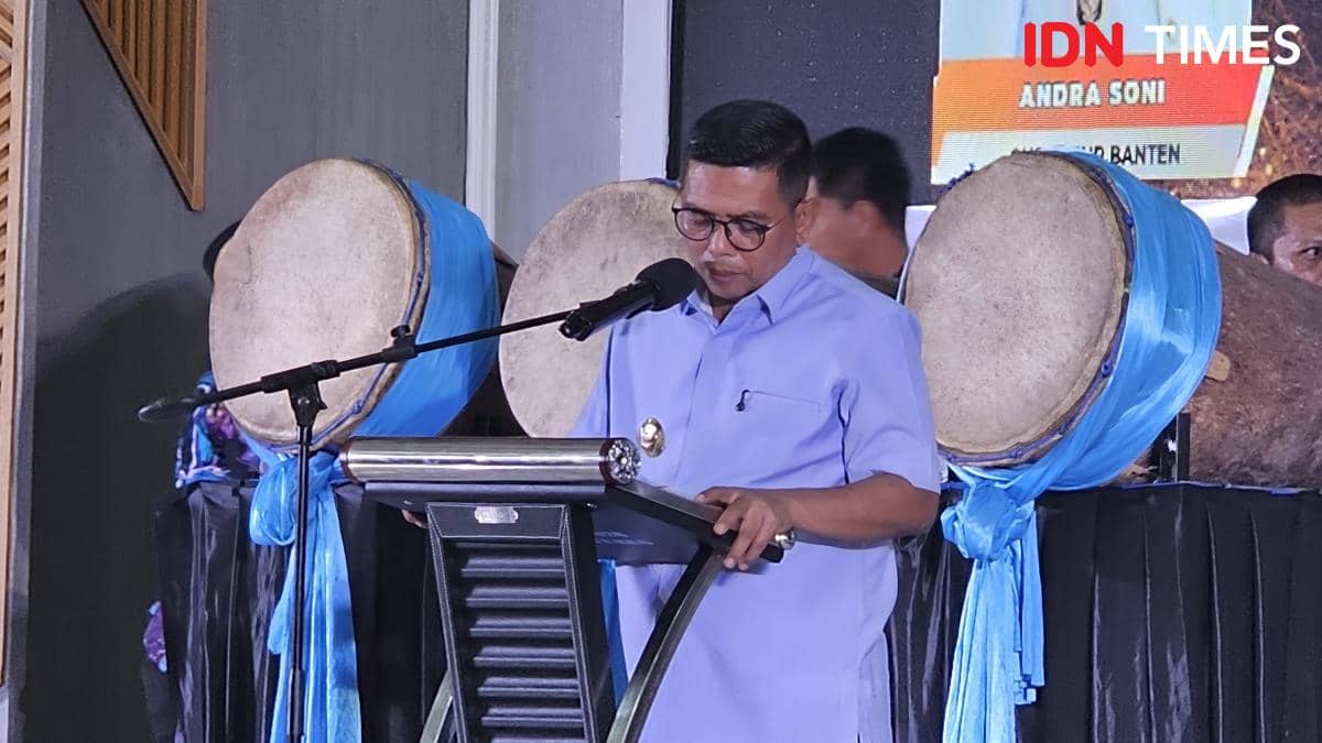 Seorang pria mengenakan kemeja biru muda berbicara di podium dengan mikrofon, di belakangnya terdapat alat musik bedug besar berhias kain biru.