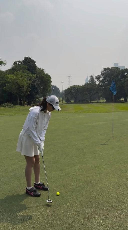 Seorang perempuan mengenakan pakaian golf putih sedang bersiap memukul bola di lapangan golf dengan latar pepohonan hijau.