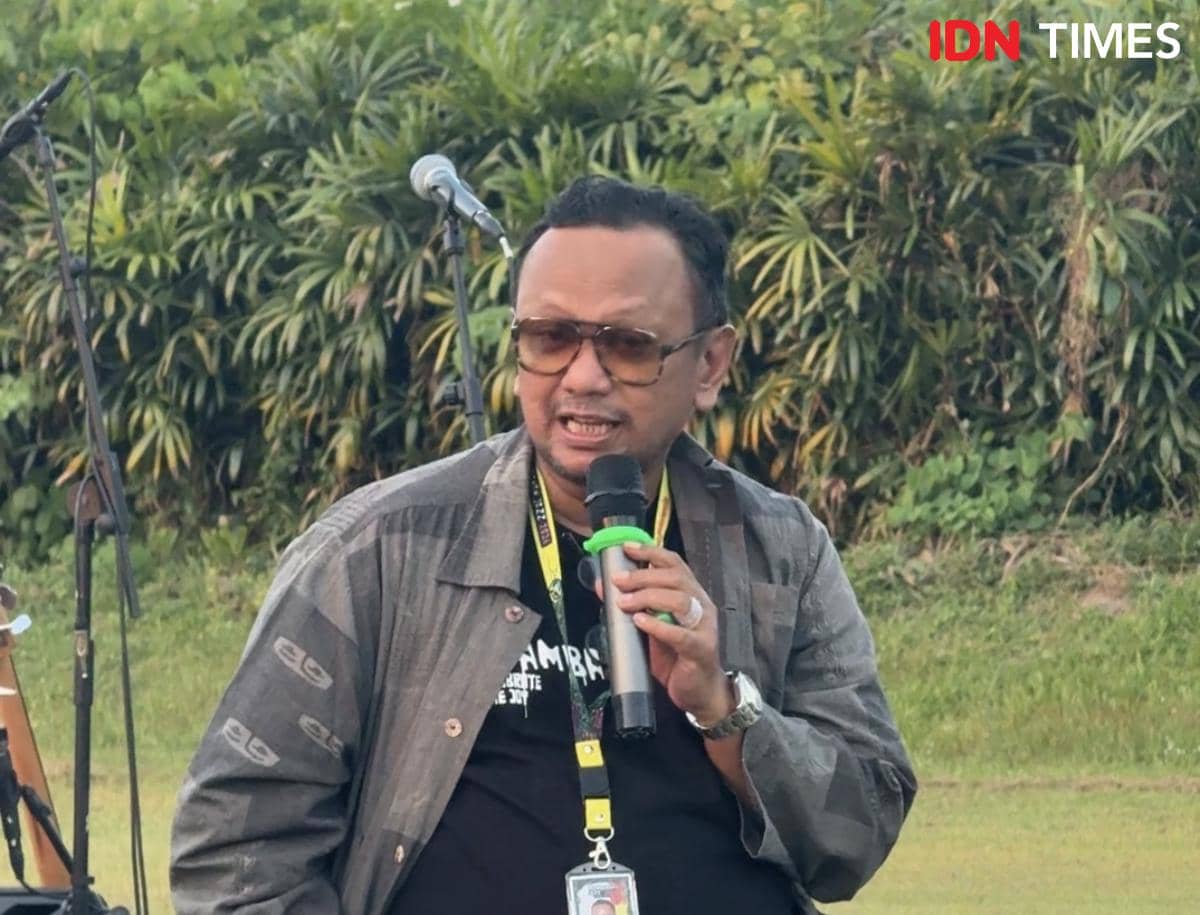 Seorang pria mengenakan kacamata dan jaket abu-abu berbicara menggunakan mikrofon di area luar ruangan dengan latar pepohonan.