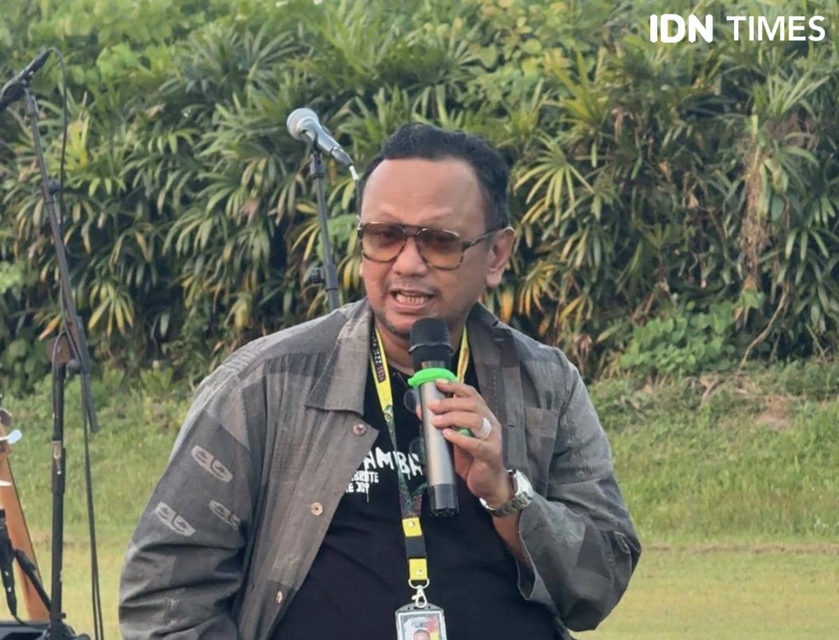 Seorang pria mengenakan kacamata dan jaket abu-abu berbicara menggunakan mikrofon di area luar ruangan dengan latar pepohonan.