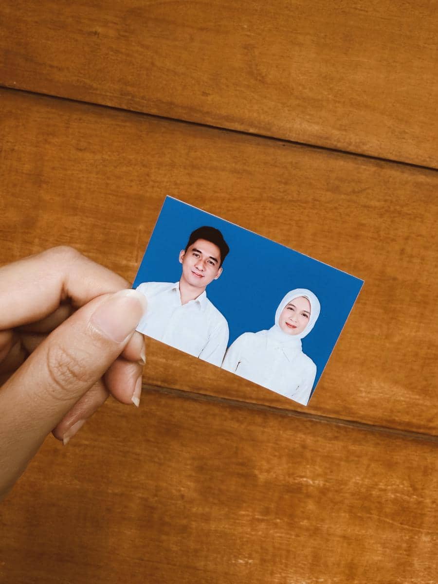 Seseorang memegang foto pasangan pria dan wanita berpakaian putih dengan latar belakang biru di atas permukaan kayu.