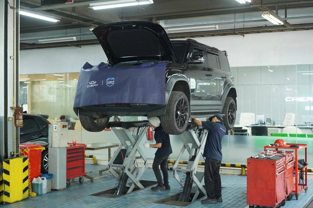 Dua teknisi melakukan perawatan mobil Chery J6 yang terangkat di bengkel resmi dengan peralatan servis lengkap.