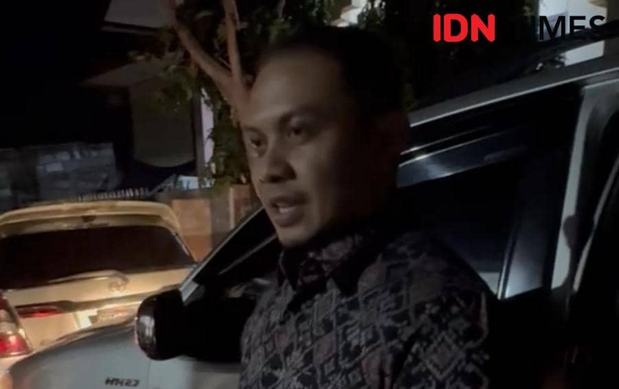 Seorang pria mengenakan batik gelap berdiri di samping mobil pada malam hari dengan latar area parkir yang remang.