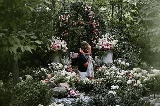 Seorang pria berlutut melamar seorang wanita di taman penuh bunga berwarna-warni dengan latar pepohonan hijau yang rimbun.