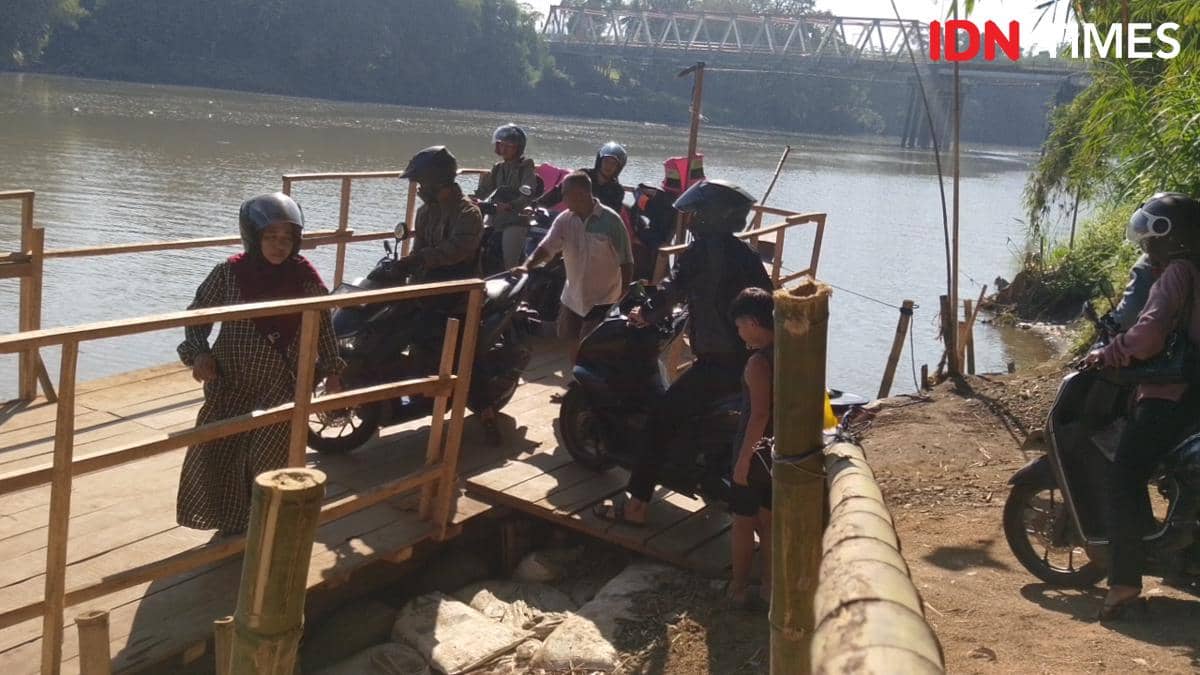Sejumlah warga bersama pengendara motor menunggu giliran menyeberang menggunakan rakit kayu di Sungai Serayu pada siang hari.