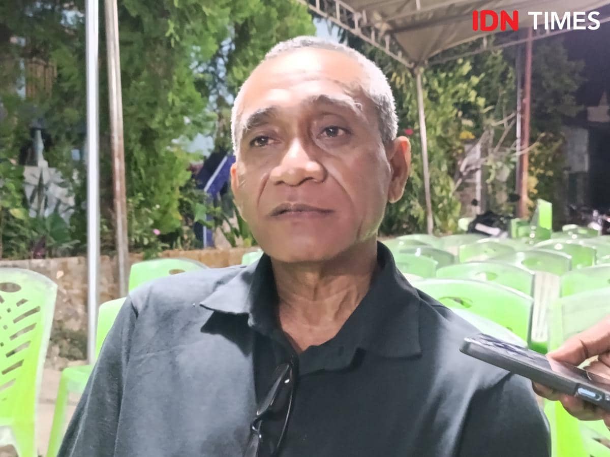 Seorang pria berambut pendek mengenakan kemeja hitam sedang berbicara di area dengan kursi plastik hijau di rumah duka.