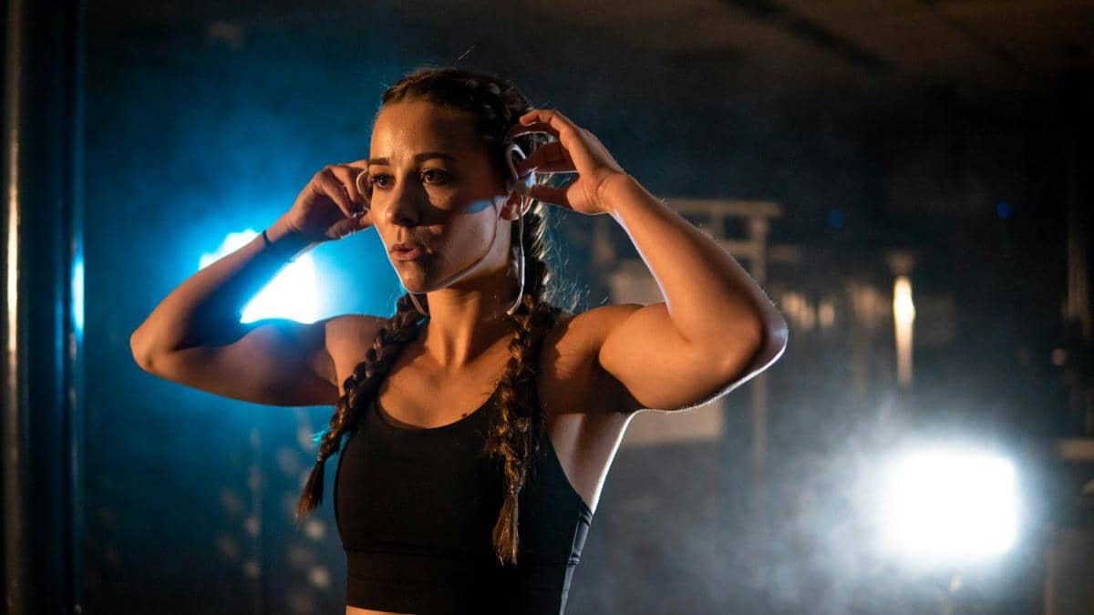 Seorang wanita berolahraga mengenakan bone conduction earphone di telinga dengan latar pencahayaan biru dan suasana gym.