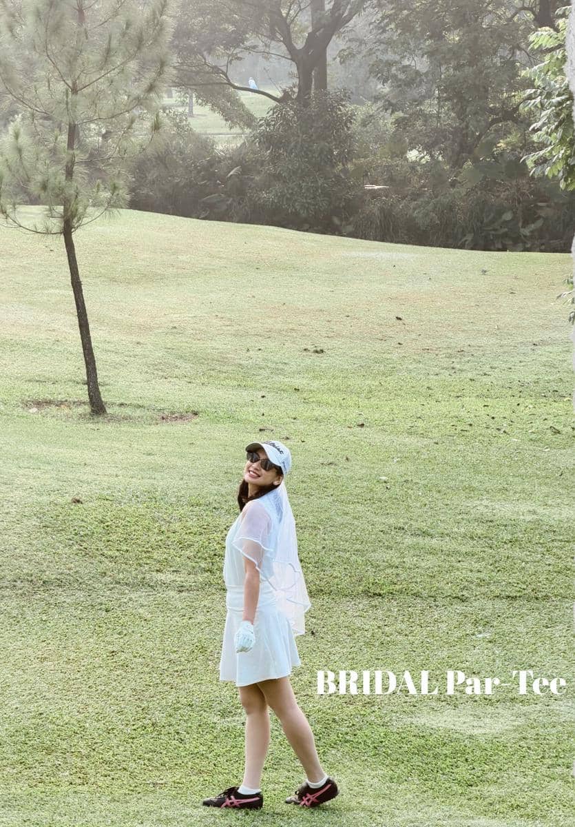 Seorang perempuan mengenakan gaun putih dan kerudung pendek bergaya pengantin berdiri di lapangan hijau dengan latar pepohonan.