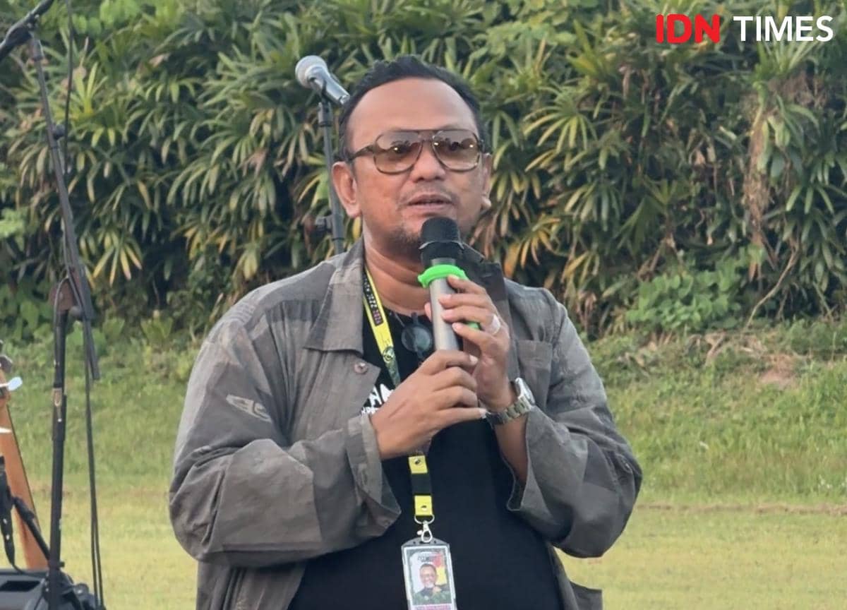 Seorang pria mengenakan jaket abu-abu dan kacamata berbicara menggunakan mikrofon di area luar ruangan dengan latar pepohonan.