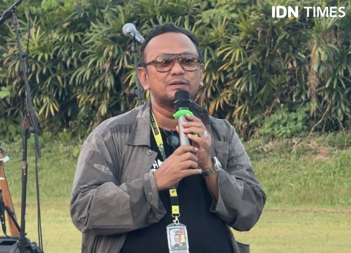 Seorang pria mengenakan jaket abu-abu dan kacamata berbicara menggunakan mikrofon di area luar ruangan dengan latar pepohonan.