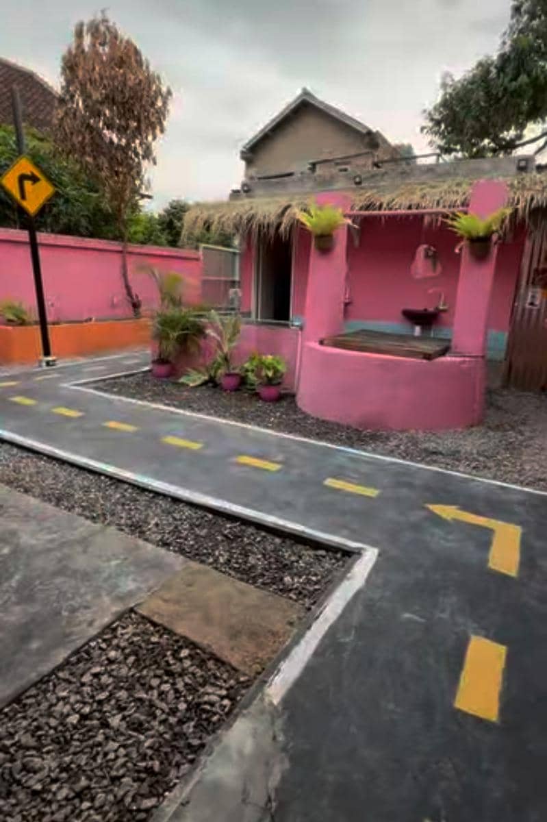 Rumah berwarna pink dengan atap jerami dan taman kecil di depan, terdapat jalan berpola marka kuning menyerupai jalan raya.