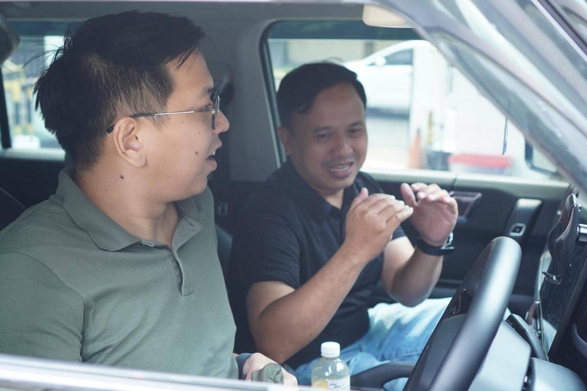Dua pria duduk di dalam mobil sedang berdiskusi, salah satunya mengenakan kaus hijau dan yang lain kaus hitam di kursi pengemudi.