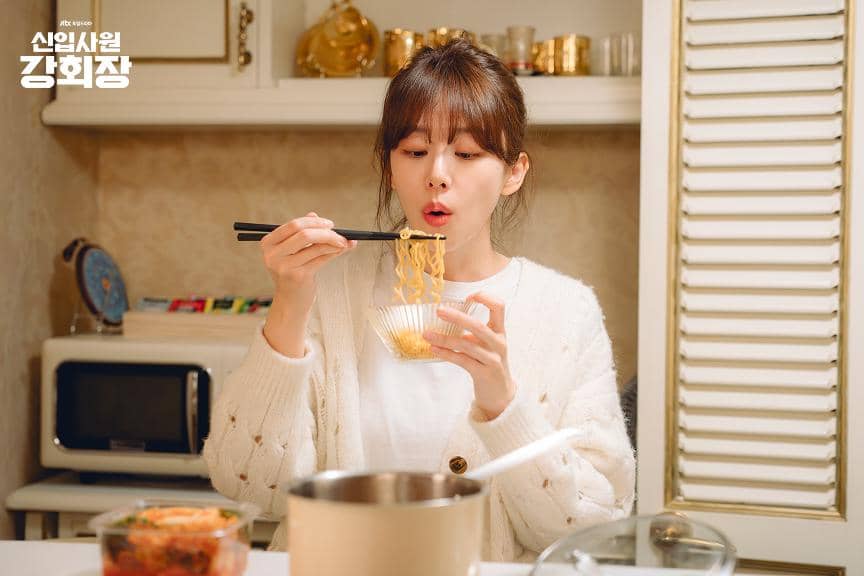 Seorang wanita mengenakan kardigan putih sedang meniup mie panas dengan sumpit di dapur yang hangat dan rapi.