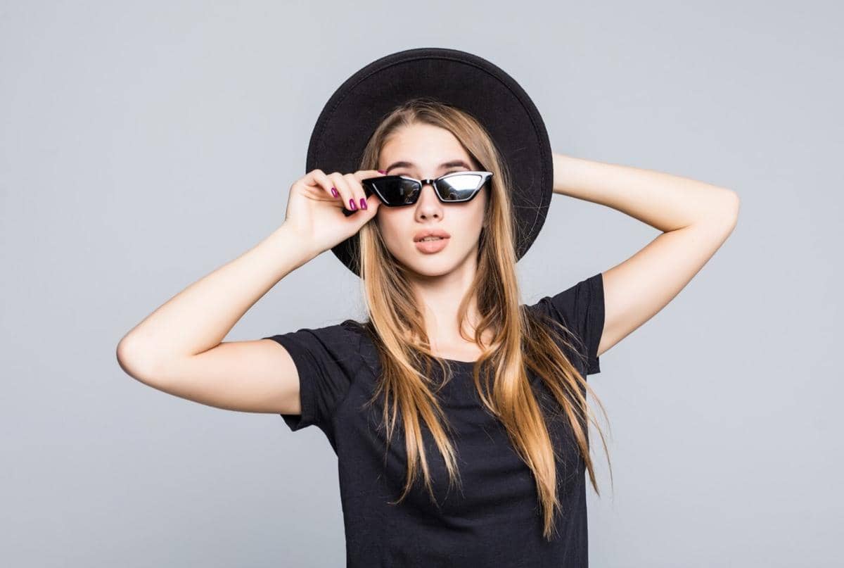 Seorang perempuan berambut panjang mengenakan topi hitam dan kacamata hitam bergaya modern dengan latar abu-abu polos.