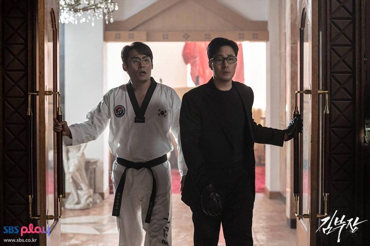 Dua pria membuka pintu besar, satu mengenakan seragam taekwondo dan satu berpakaian hitam dalam adegan drama Korea Agent Kim Reactivated.