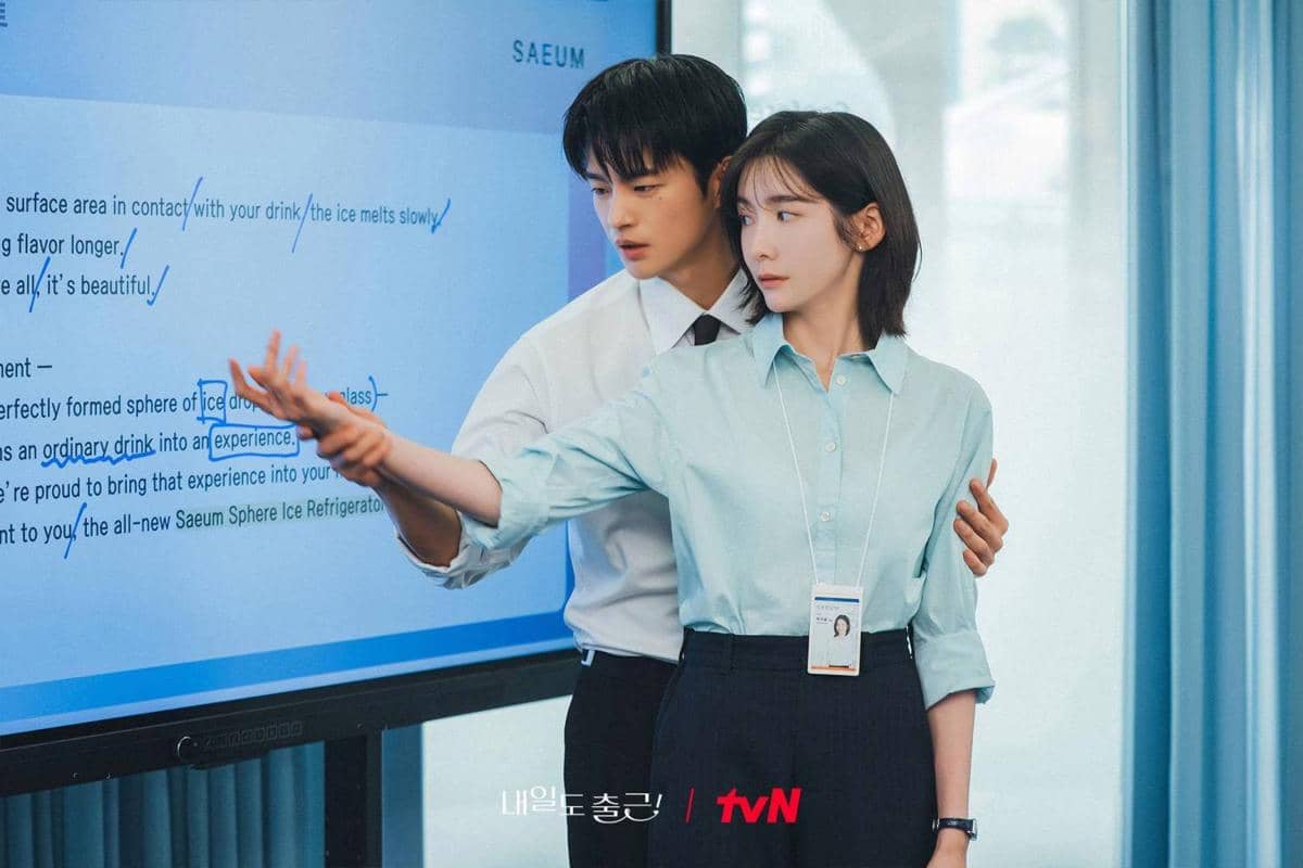 Dua karyawan berdiri di depan layar presentasi, salah satunya membantu menunjukkan sesuatu di layar dalam adegan drama Korea See You at Work Tomorrow.