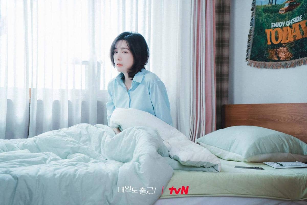 Seorang wanita berambut pendek duduk di tempat tidur dengan selimut putih di kamar bercahaya lembut, mengenakan kemeja biru muda.
