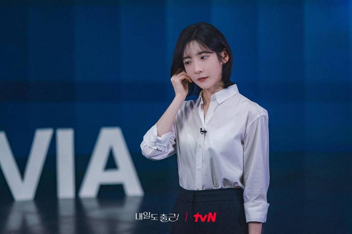 Seorang wanita berambut pendek mengenakan kemeja putih dan rok hitam berdiri di depan latar biru dengan logo tvN di bawahnya.
