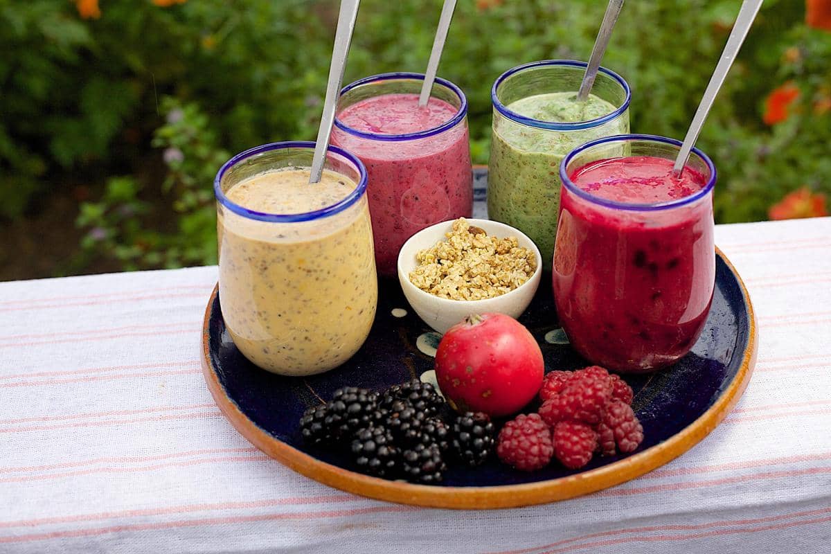 Ilustrasi dua gelas smoothie buah berwarna kuning dan merah muda, dikelilingi stroberi, kiwi, pisang, dan buah segar.