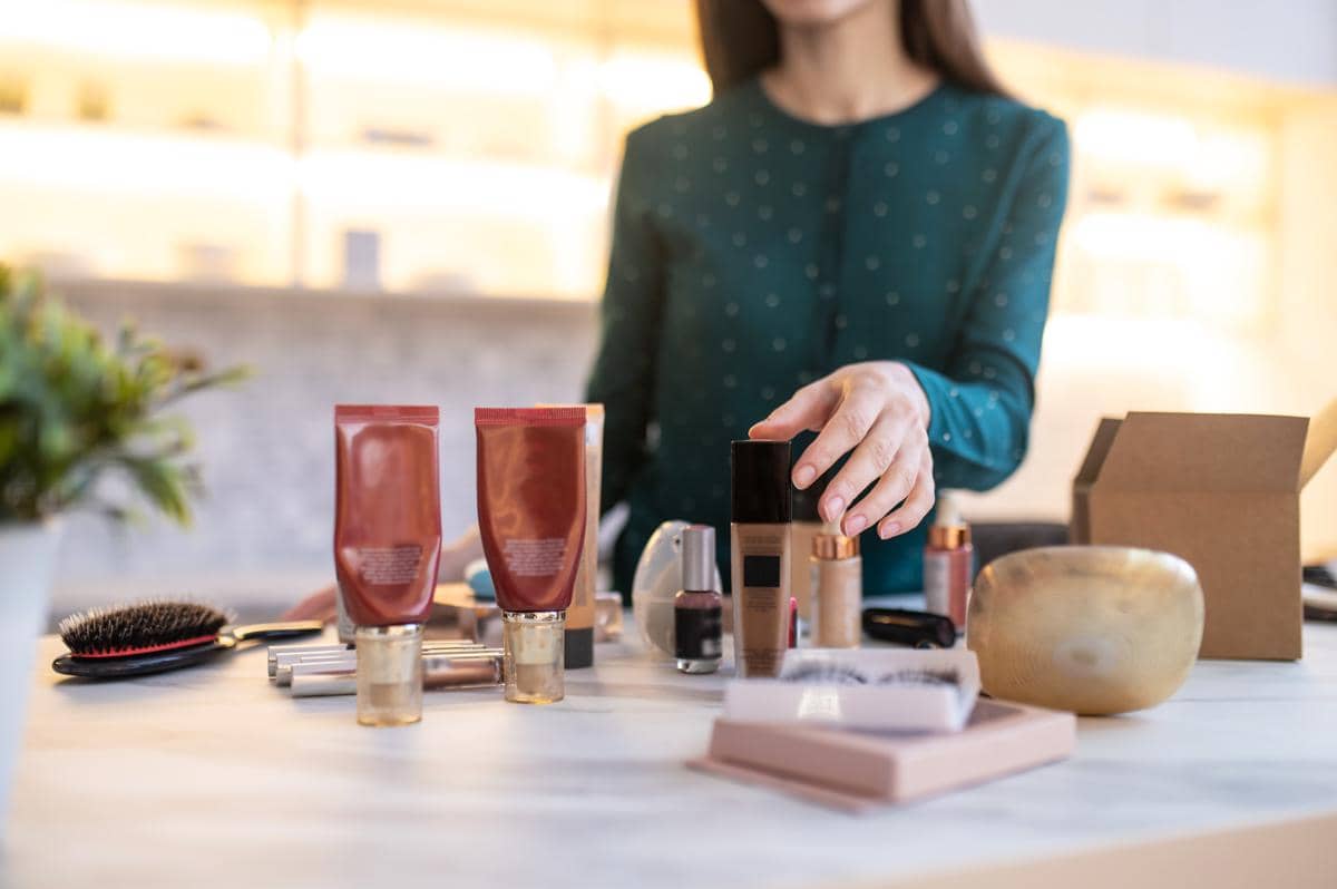Seorang wanita mengenakan blus hijau sedang merapikan berbagai produk kosmetik seperti foundation, lipstik, dan krim di atas meja.