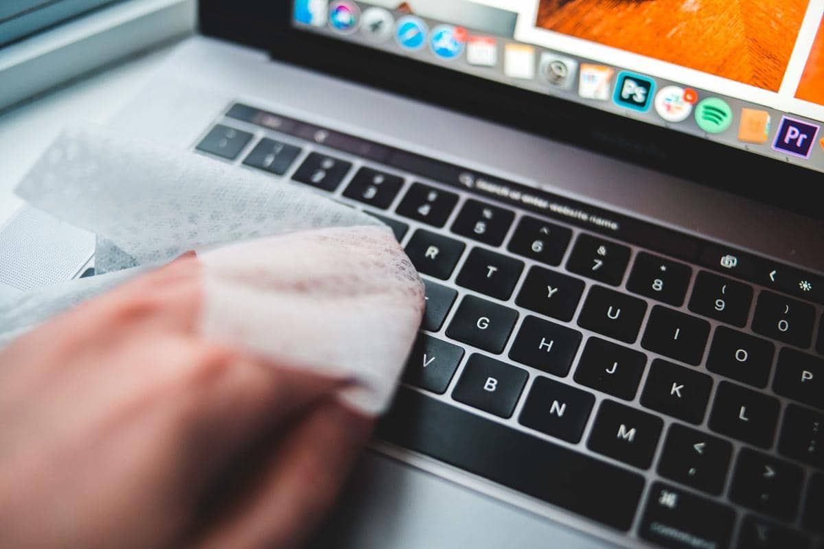 Tangan sedang mengelap keyboard laptop dengan tisu lembap untuk membersihkan permukaan tombol dari debu dan kotoran.