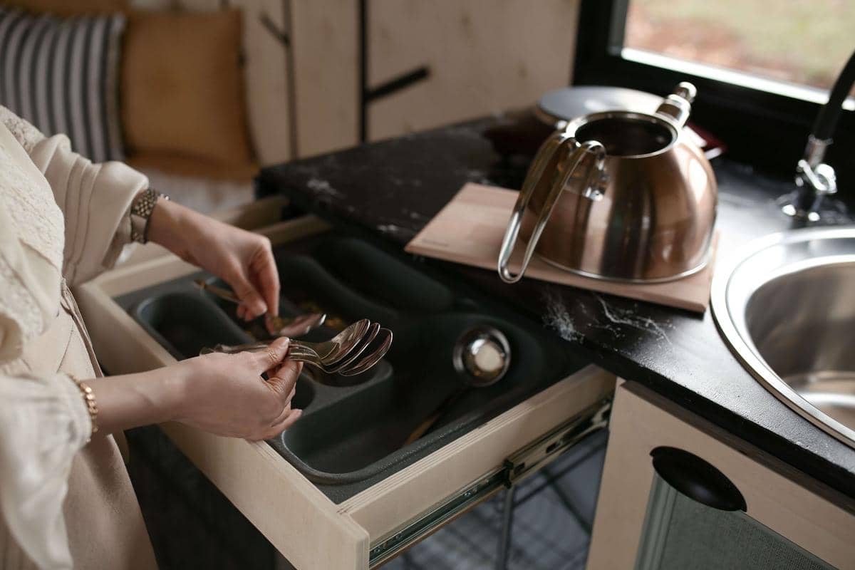 Seseorang sedang merapikan sendok dan garpu ke dalam laci dapur di dekat wastafel dan teko logam di atas meja hitam.