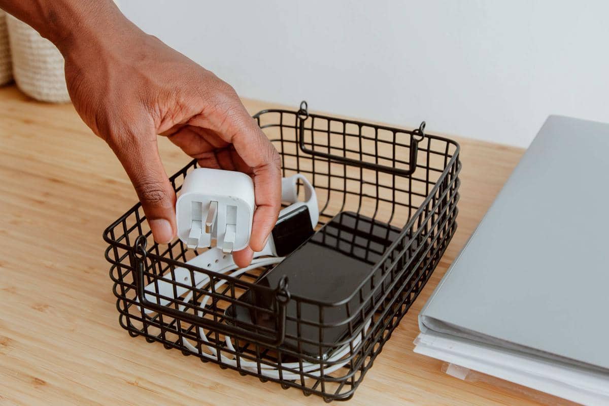 Tangan seseorang menata adaptor dan perangkat elektronik kecil seperti ponsel serta kabel di dalam keranjang kawat hitam di atas meja kayu.