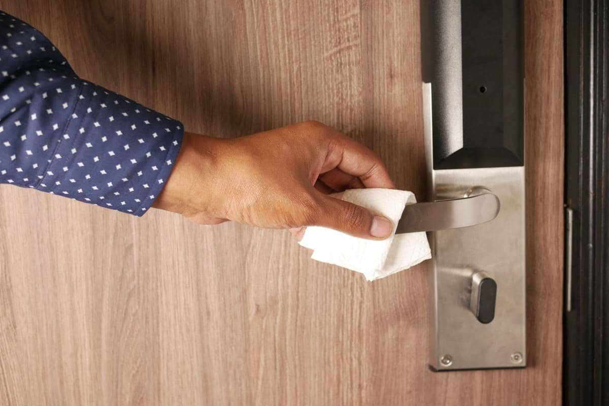Tangan seseorang mengenakan kemeja biru sedang mengelap gagang pintu logam dengan tisu di permukaan pintu kayu.