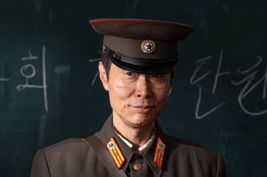 Seorang aktor mengenakan seragam militer lengkap dengan topi dan lambang, berdiri di depan papan tulis dengan tulisan Korea di latar belakang.