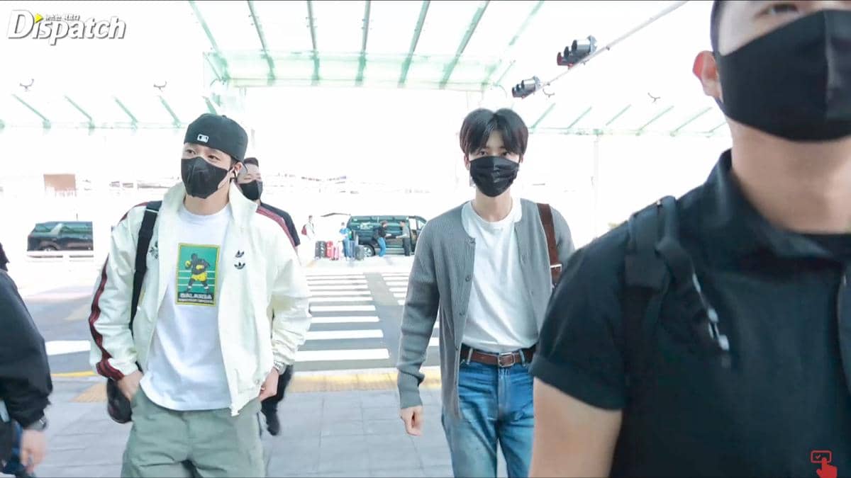 Dua pria mengenakan masker hitam berjalan di area keberangkatan bandara dengan pakaian kasual dan membawa tas.