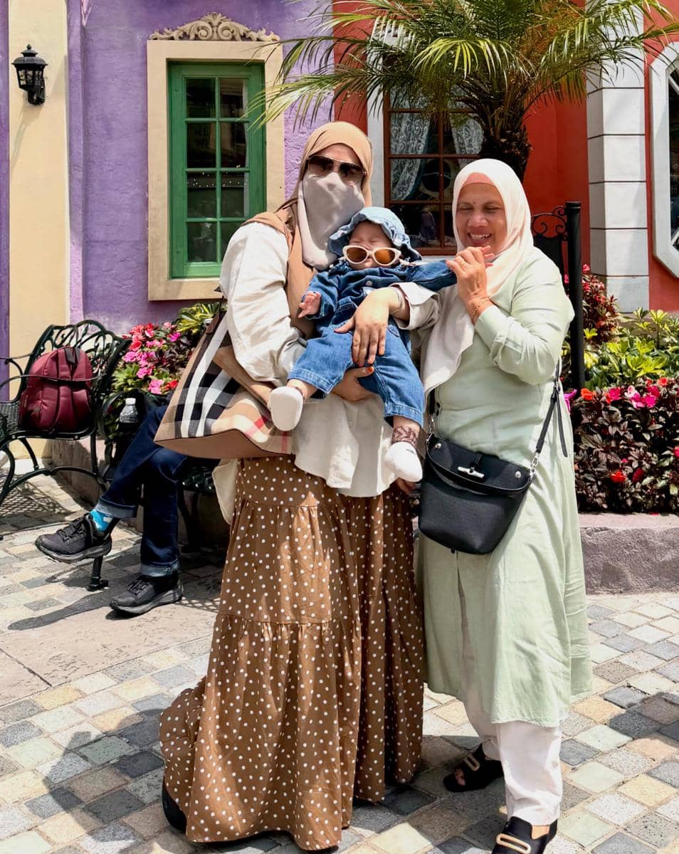 Dua wanita berhijab berpose bersama seorang bayi di luar ruangan dengan latar bangunan berwarna ungu dan oranye di Malaysia.