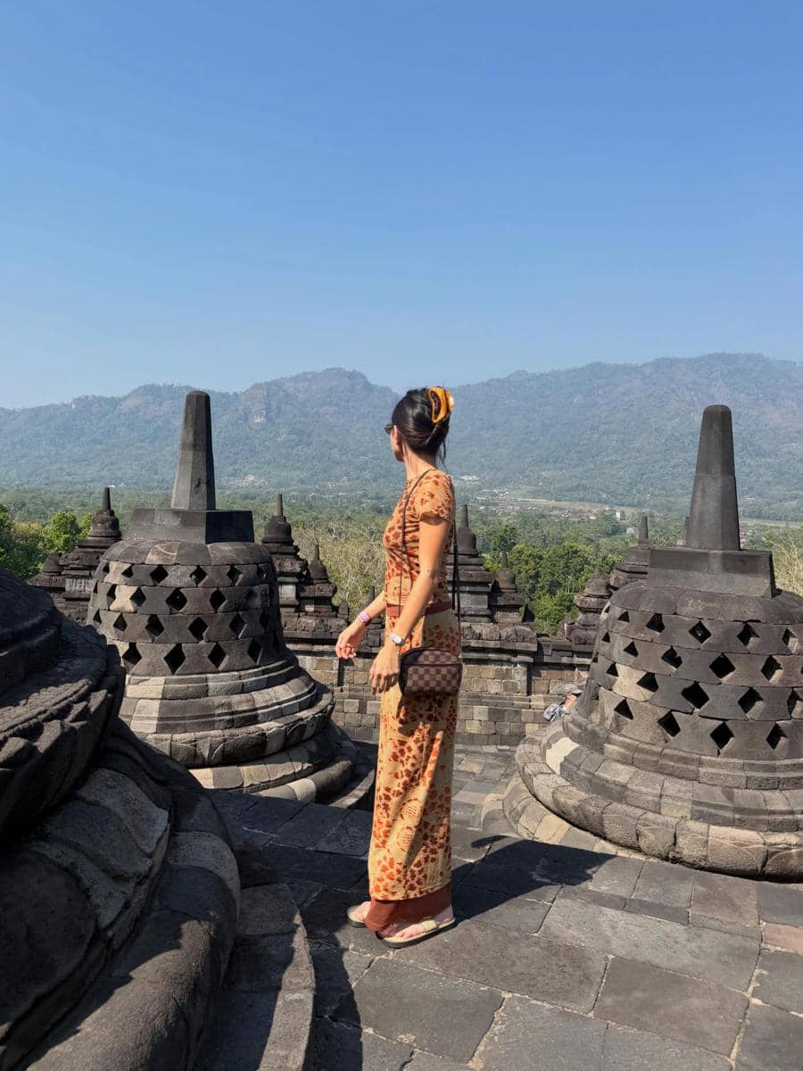 Seorang perempuan mengenakan dress bermotif batik berdiri di antara stupa Candi Borobudur dengan latar pegunungan dan langit biru cerah.