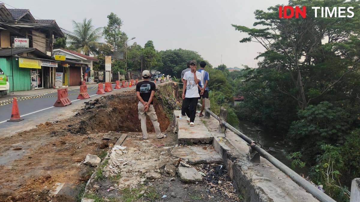 Beberapa orang berdiri di tepi jalan yang longsor di Jl Kebon Pedes, dengan area jalan rusak dan kerucut lalu lintas di sekitarnya.
