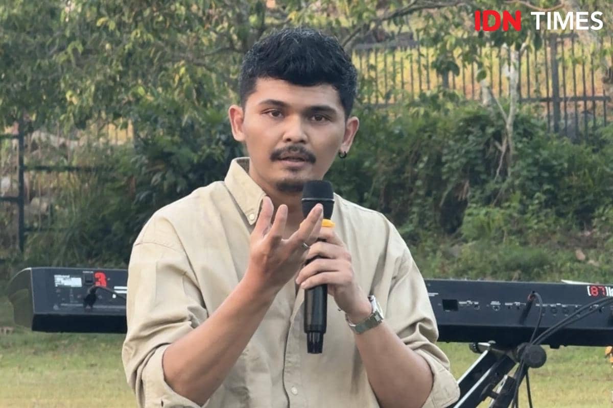 Seorang pria mengenakan kemeja krem memegang mikrofon saat berbicara di depan keyboard dalam konferensi pers luar ruangan