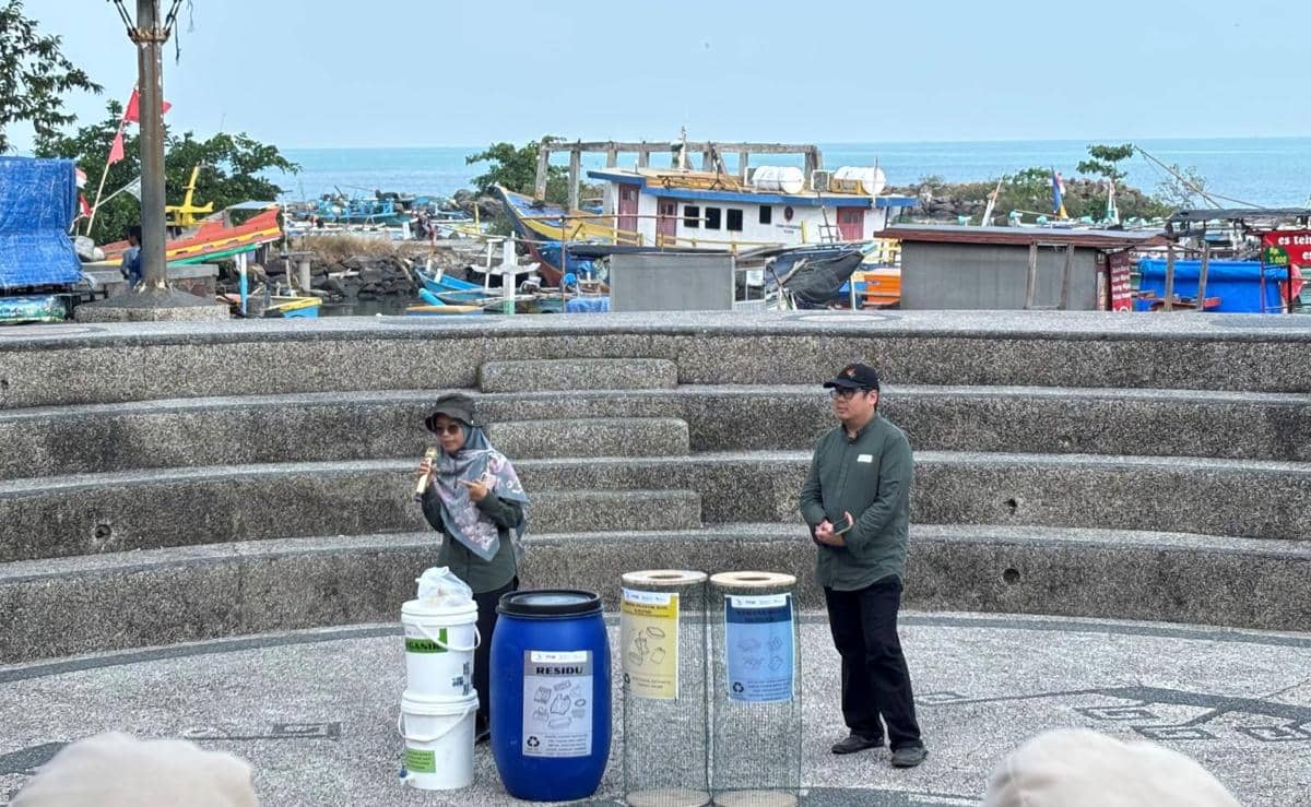 Dua peserta berdiri di area terbuka dekat dermaga dengan latar perahu nelayan, menjelaskan pengelolaan sampah dalam program wisata berkelanjutan.
