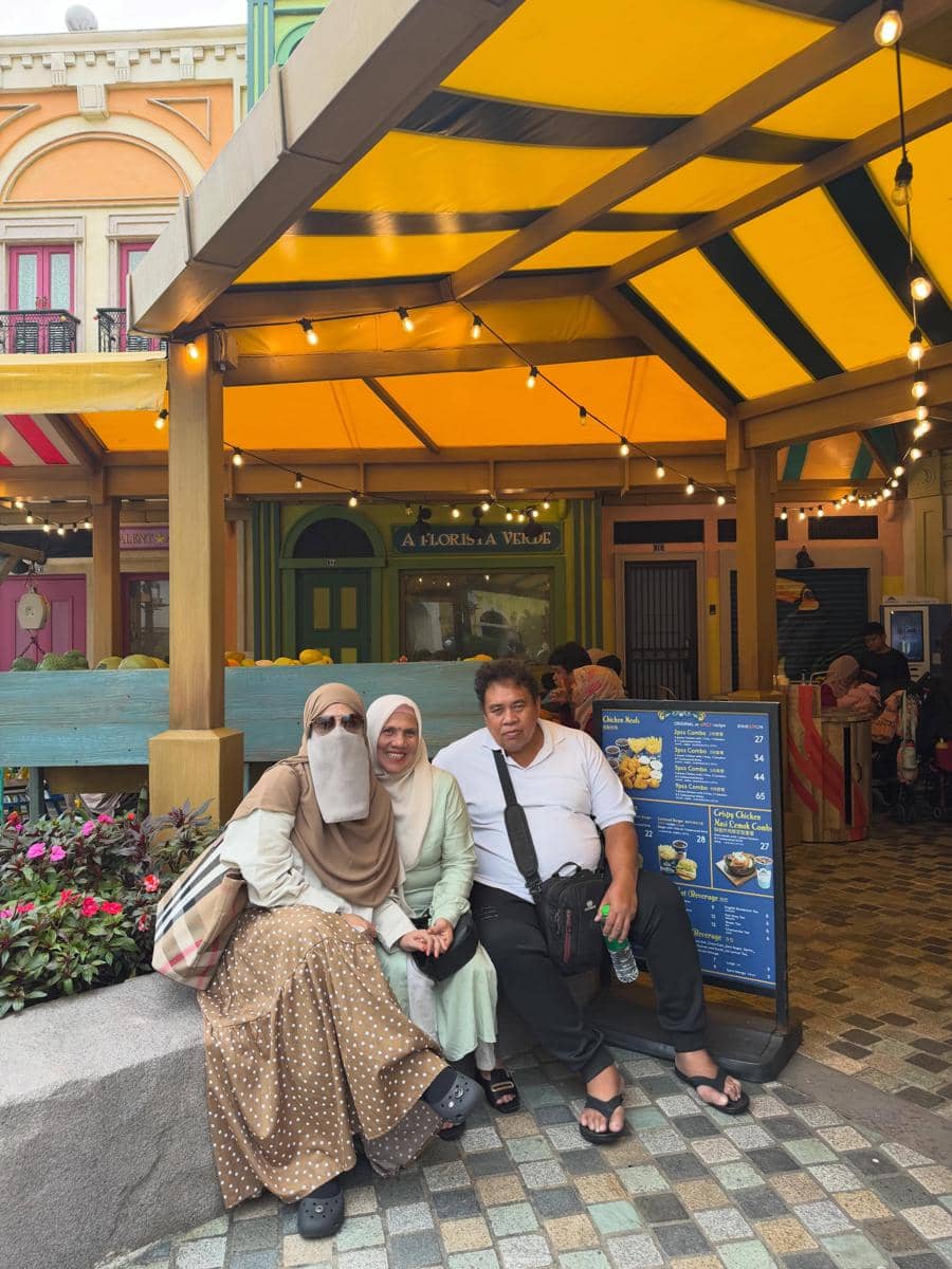 Tiga orang duduk bersama di area outdoor berwarna cerah dengan atap kuning dan lampu gantung di Malaysia.